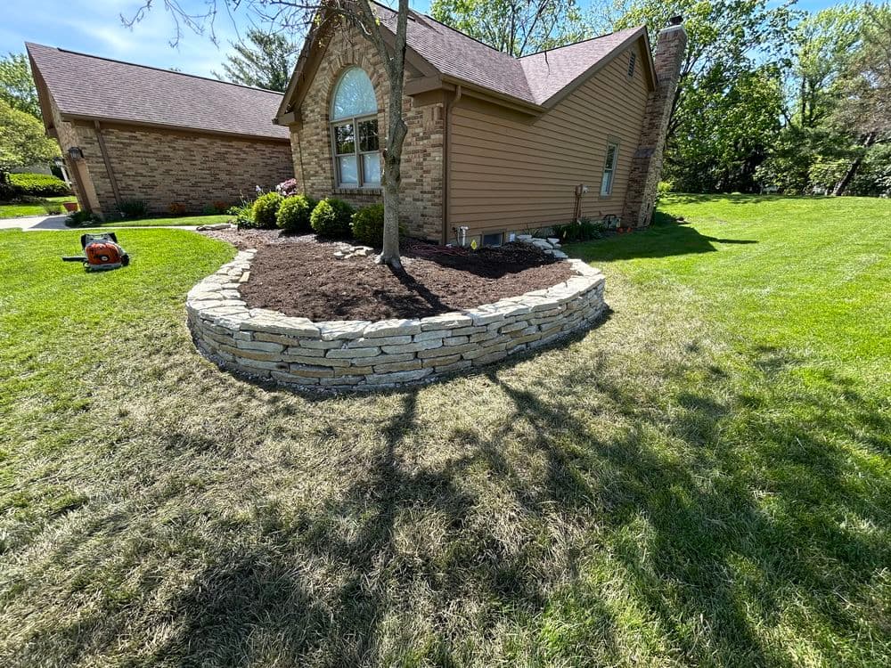 Stone landscaping border around a tree and mulch bed next to a house in a green yard.