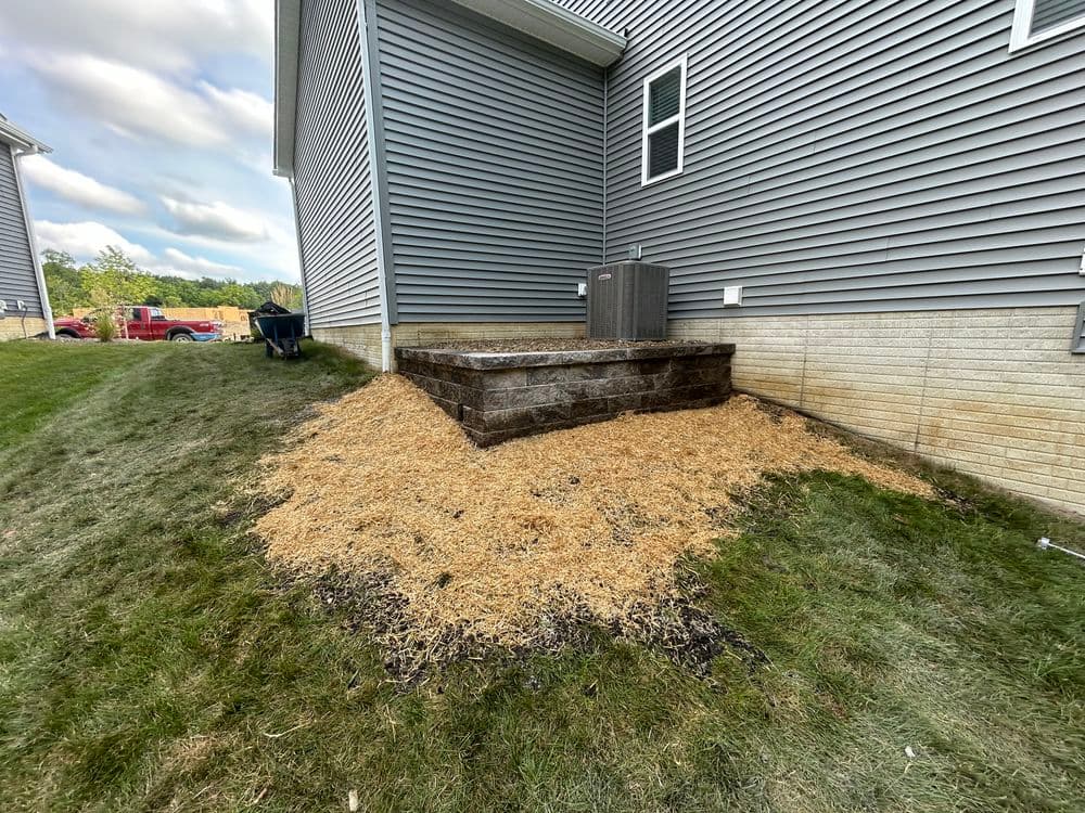 Landscaped area with a stone flower bed and mulch near a house's side. Green grass and sky visible.