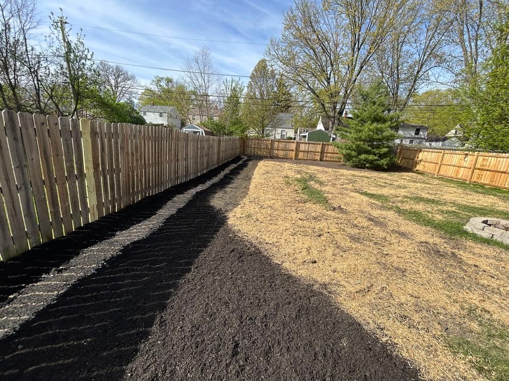 Landscaped backyard with freshly tilled soil, wooden fence, and greenery under a blue sky.