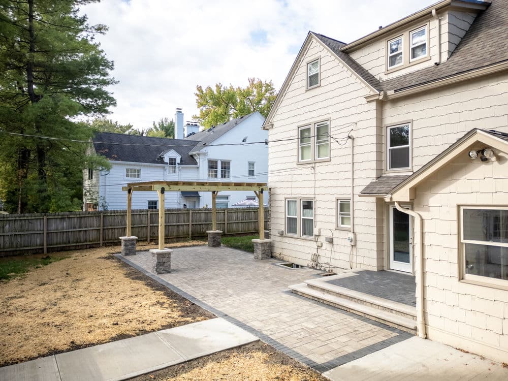 Backyard view of a house with a stone patio, pergola, and landscaped area.