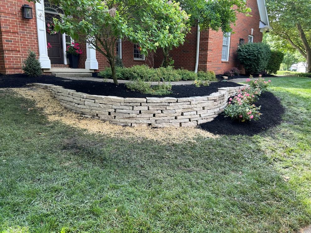 Landscaped garden with stone retaining wall, black mulch, and blooming flowers.