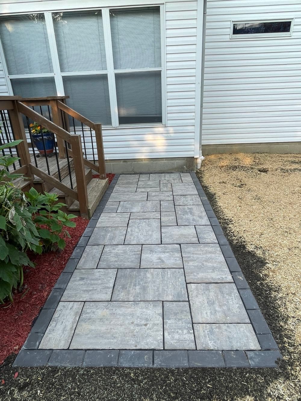 Gray patio stone walkway beside a house, bordered by black edging and red mulch.