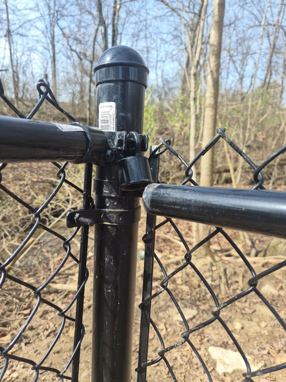 Black chain-link fence post and gate latch against a backdrop of trees and blue sky.