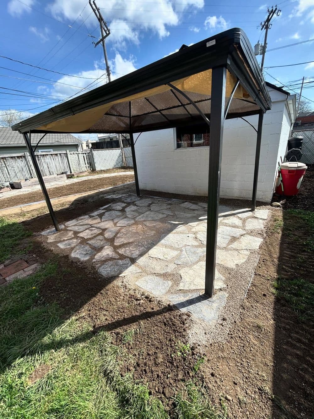 Outdoor stone patio with a metal canopy in a residential backyard on a sunny day.