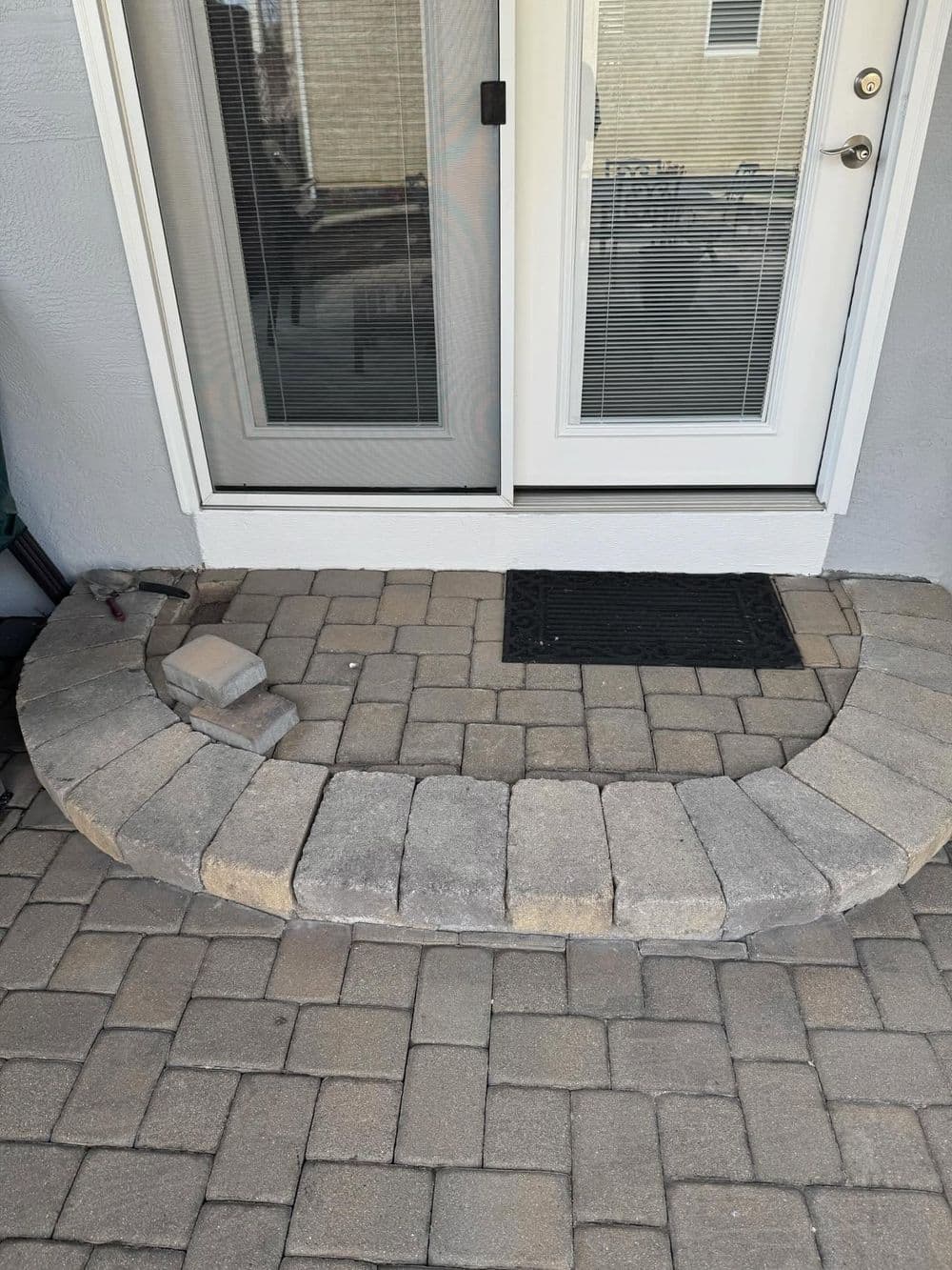 Curved stone entryway with pavers leading to glass door, featuring a black mat.